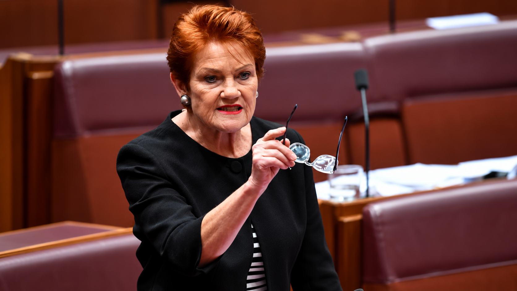 Pauline Hanson speaking in the Australian Senate chamber, holding her glasses while addressing Parliament.